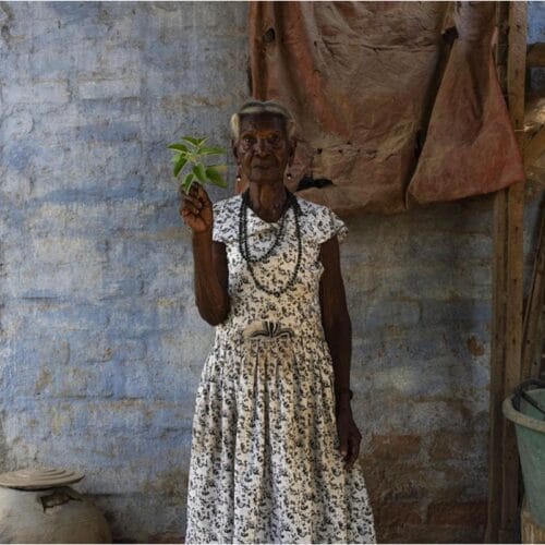 Retrato de una mujer mayor con vestido estampado frente a una pared azul, con una rama verde en la mano. Proyecto Rosalina, de Joana Toro.