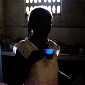 Niña a contraluz sosteniendo una taza azul en una cocina, re Niña a contraluz sosteniendo una taza azul en una cocina, recortada contra la luz de una ventana. Fotografía de Joana Toro.