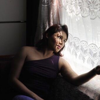 Mujer sentada junto a una ventana con visillo de encaje, ilu Mujer sentada junto a una ventana con visillo de encaje, iluminada por la luz lateral. Fotografía de Joana Toro.